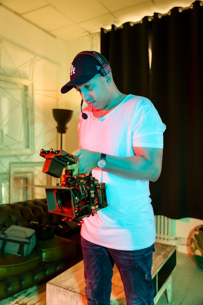 A cameraman in a white t-shirt with headset operates a professional video camera indoors.