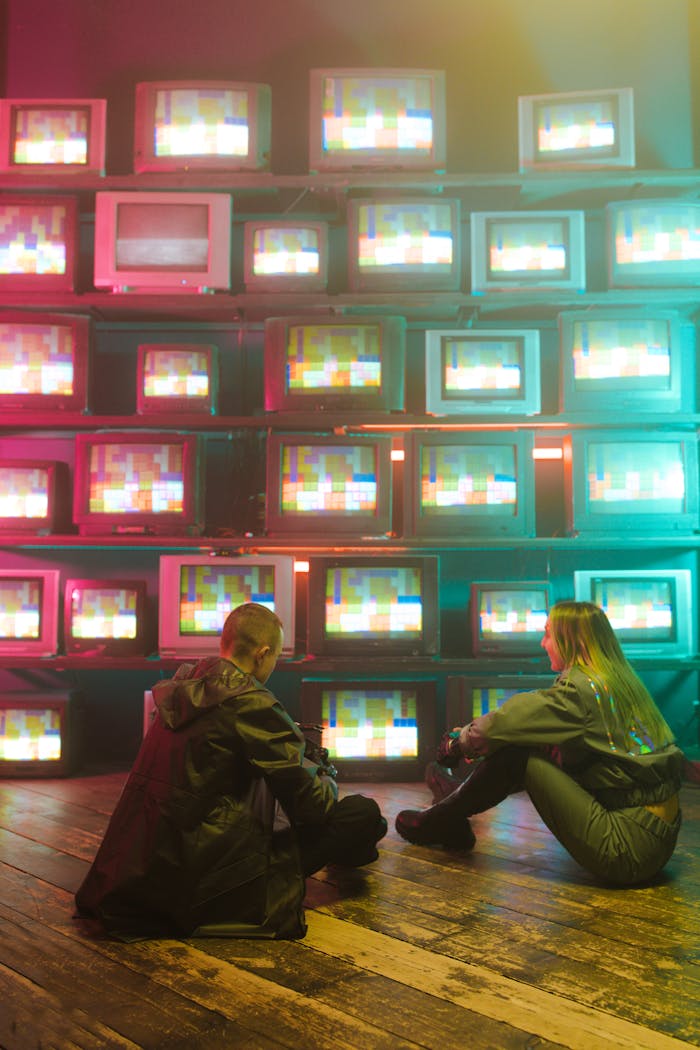 Two gamers in futuristic attire sit by a neon-lit wall of retro TVs.